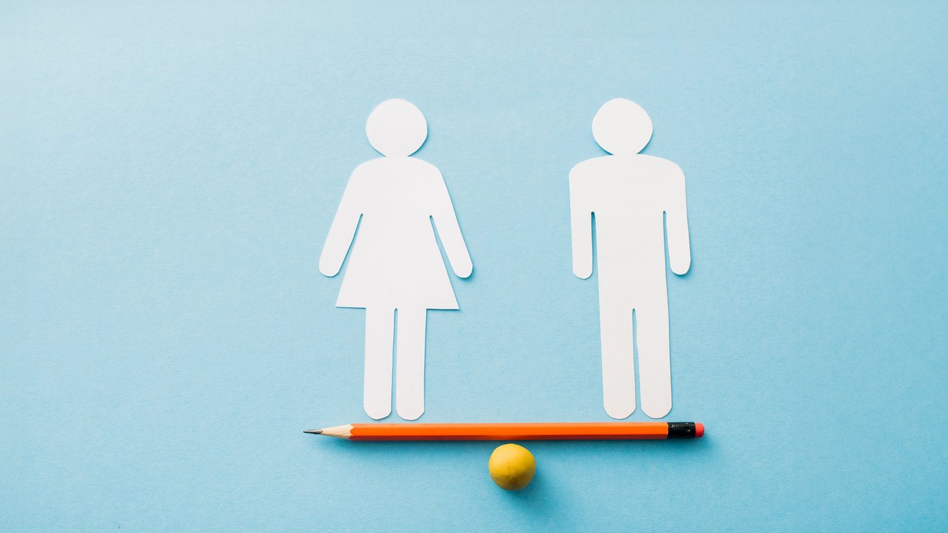 The image shows a man and a woman in paper cut-out standing at each end of a pencil that serves as a seesaw. The seesaw is balanced.